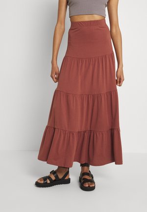 Femme portant une jupe maxi taille haute à volants couleur rouille, des sandales noires à brides et un top court taupe.