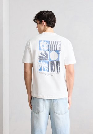 Homme de dos portant un t-shirt blanc avec un imprimé graphique floral et géométrique bleu et un jean bleu clair, sur fond uni.