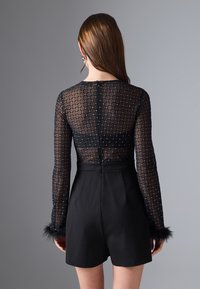 Bodysuit negro con largas mangas transparentes que presentan un patrón de diamantes y acentos plateados, combinado con unos pantalones cortos negros de talle alto.