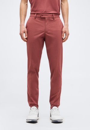 Muž, ktorý má na sebe úzke tehlovo červené nohavice a biele tenisky, stojí na jednoduchom svetlosivom pozadí.