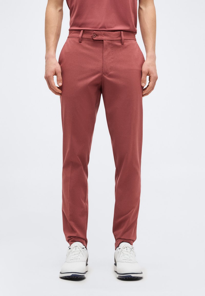 Άνδρας που φοράει εφαρμοστά βερμούδα σε τούβλινο κόκκινο χρώμα και λευκά αθλητικά παπούτσια, στέκεται μπροστά σε ένα απλό ανοιχτό γκρι φόντο.