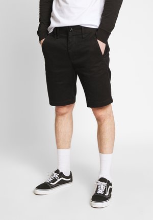 Shorts en coton noirs à coupe droite, avec poches latérales et fermeture à bouton. Associés à des chaussettes blanches et des baskets noires.