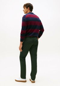 Jonge man staat, draagt een marineblauwe en bordeauxrode gestreepte trui, donkergroene broek en witte sneakers met gumrubberen zolen, gericht naar links.