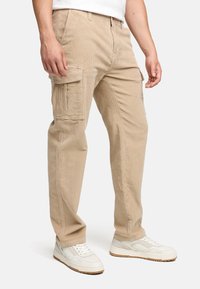 Beige kordflorsbyxor med sidofickor, raka ben och synlig textur. Bärs med vita sneakers mot en neutral bakgrund.