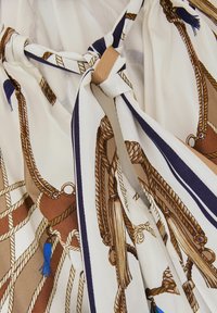 Detailní pohled na krémovou látku s potiskem hnědého lana a střapců, obsahující uvázaný uzel s modrobíle pruhovanými okraji.