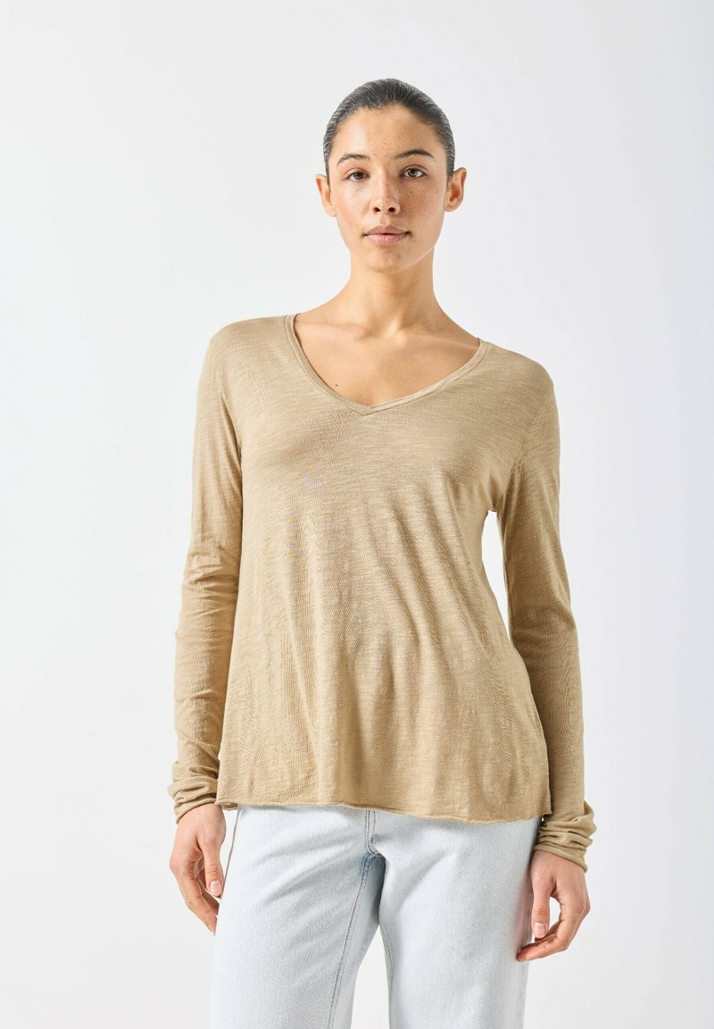 Top beige de manga larga con escote en V, hecho de tela ligera y texturizada, combinado con vaqueros azul claro. Corte simple y relajado con un brillo sutil.