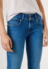 Nærbilde av en person som har på seg blå høyt midje denimjeans med dobbel knappelukking og en hvit skjorte som er stukket inn.