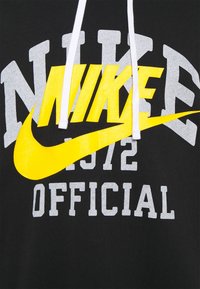 Svart sweatshirt med en stor, gul Nike-logotyp, grå text som säger "NIKE 1972 OFFICIELL", med vita snoddar vid huven.