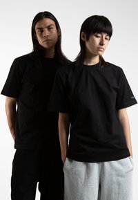 To personer står tæt sammen, begge iført sorte t-shirts, mens kvinden har grå sweatpants på. Den neutrale baggrund fremhæver fokus på tøjet.