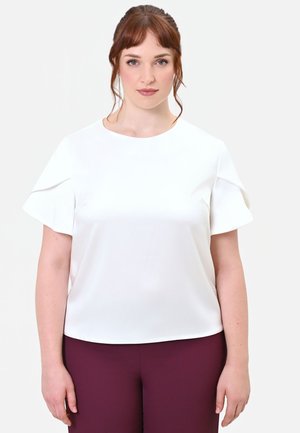 Donna con capelli rossi che indossa una camicetta bianca a maniche corte con maniche arruffate e pantaloni viola scuro in piedi contro uno sfondo semplice.