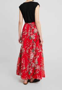 Robe maxi avec un haut à manches courtes noires et une jupe florale rouge ornée de fleurs roses et de feuilles vertes. Tissu léger et fluide.