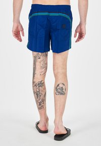 Costume da bagno blu con strisce verdi e nere, tasca laterale, cintura elasticizzata e tessuto leggero. Indossato con ciabatte nere.