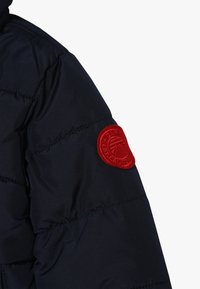 Marinblå pufferjacka med kviltat mönster och en cirkulär röd logopatch på överarmen. Materialet är slätt och lätt glansigt.