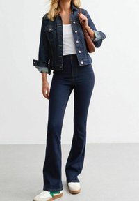 Chaqueta de denim azul oscuro con botones al frente y mangas dobladas, combinada con jeans acampanados de cintura alta en azul oscuro y zapatillas blancas con detallitos verdes.