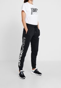 Vit T-shirt med svart grafisk text på framsidan, matchad med svarta joggers med en vit logotyp. Svarta sneakers fulländar outfiten.