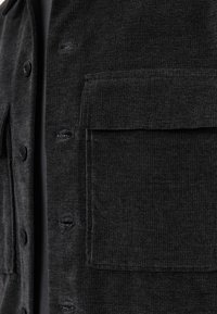 Black textured shirt jacket with a button closure, featuring a large front pocket and visible stitching details. Crafted from a soft, woven fabric.