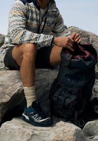 Homme en polaire à motifs et short assis sur des rochers, ajustant un sac à dos de randonnée noir, portant des chaussettes beige et des chaussures de randonnée bleues.