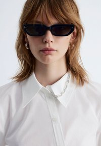 Black rectangular sunglasses with dark lenses, worn with a white collared shirt. Silver chain necklace and small earrings visible.