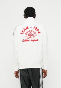 Weißes Sweatshirt mit Reißverschluss, roter Blumenstickerei, dem Schriftzug "TEAM - 1994" und dem "Adidas Originals" Logo auf der Rückseite; mit geripptem Kragen und Bündchen.