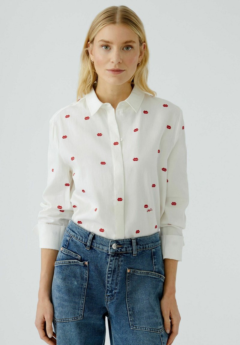 White button-up shirt featuring red lip embroidery, long sleeves, and a classic collar, paired with high-waisted blue denim jeans.