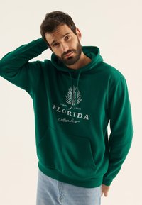 Sudadera verde hecha de tela suave, con un bolsillo tipo canguro, texto blanco con "Florida" y un gráfico de una palmera. Puños y dobladillo acanalados.