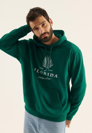 Hoodie vert en tissu doux, comprenant une poche kangourou, un texte blanc avec "Florida" et un graphique de palmier. Poignets et ourlet côtelés.