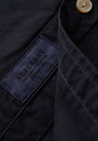 Donkerblauwe shirtkraag met een zichtbare beige knop en het gestikte Superdry merklabel aan de binnennaad.