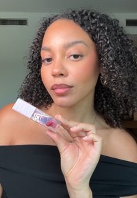 Heldere lip plumper in een rechthoekige, doorzichtige tube met een roze accent. Model toont het met krullend haar, gekleed in een zwarte off-shoulder top.