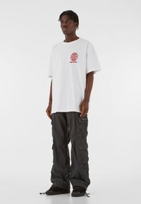 Valge liiga suur t-särk punase graafilise disainiga, koos mustade kampsunpükstega, millel on mitu taskut ja tekstuurne kangas. Mustad kingad.