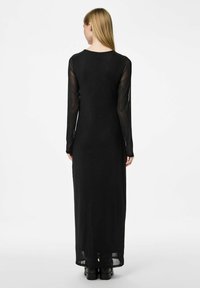 Dlouhá černá šaty s dlouhými průhlednými rukávy. Měkký materiál, přiléhavý střih a kulatý výstřih, s mírně průhledným spodním lemem.