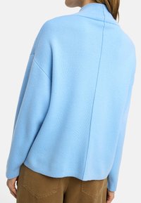 Jasnoniebieski, luźny sweter z wysokim kołnierzem, prążkowaną fakturą i opadającymi ramionami. Posiada centralny szew oraz swobodny krój.