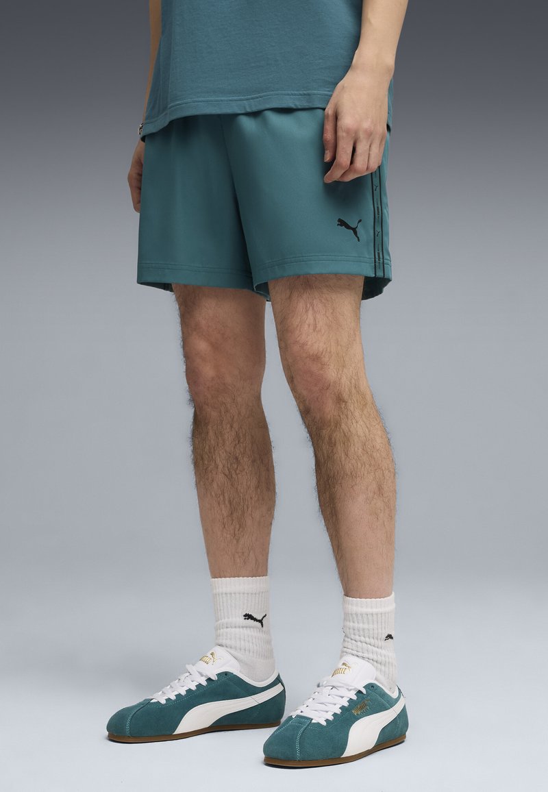 Homme portant un short Puma vert-bleu, des chaussettes blanches Puma et des baskets Puma vert-bleu, debout devant un fond gris uni.