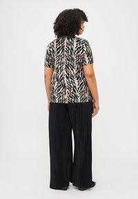 Une personne aux cheveux bouclés porte une chemise à motifs noirs et beiges et un pantalon large plissé noir, vue de dos sur fond blanc.