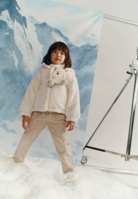 Sudadera con capucha blanca y esponjosa con detalles de oso, pantalones de pana beige, de pie sobre nieve artificial frente a un fondo montañoso.