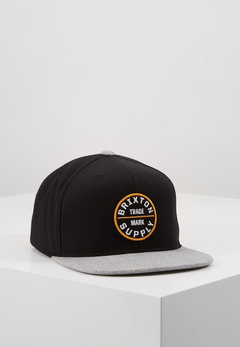 Casquette de baseball noire avec une visière plate grise et un patch circulaire avec le logo de la marque déposée Brixton Supply sur le panneau avant.
