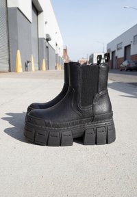 Par de botas negras de plataforma gruesas con paneles laterales elásticos, situadas en una calle de cemento en una zona industrial urbana.