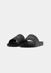 Sandales noires ouvertes à bout ouvert avec larges brides présentant le logo Calvin Klein en blanc sur un fond gris clair uni.