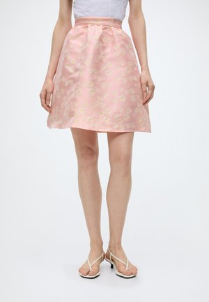 Femme portant une jupe trapèze rose clair avec de petits motifs floraux blancs et jaunes, associée à des sandales blanches à talons et un haut blanc.