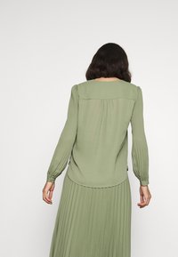 Blusa verde a maniche lunghe con design plissettato, scollatura rotonda e dettagli arricciati sulle spalle e ai polsi. Tessuto morbido e leggero.