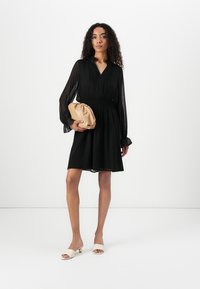 Svart kjole med lange, gjennomsiktige ermer og smocket midje; kombinert med en beige clutch og kremfargede sandaler med sklie som har en gullaccent.