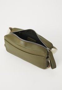 Olive green leather bag with a zippered top, a textured front pocket, and a cream-colored strap. Interior features a black lining.