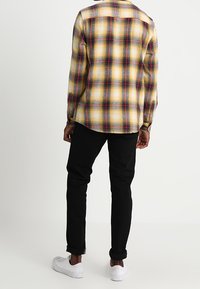 Camicia di flanella a quadri gialli, rossi e neri. Maniche lunghe, chiusura con bottoni, orlo arrotondato. Abbinata a jeans neri e sneakers bianche.