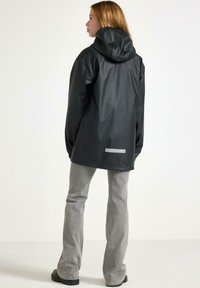 Una donna indossa un impermeabile nero con cappuccio e jeans grigi chiari, in piedi con la schiena rivolta verso uno sfondo chiaro e semplice.