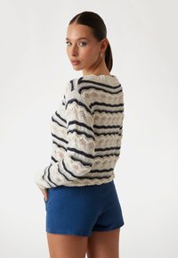 Strickpullover mit einem cremefarbenen und marineblauen Streifenmuster, das ein strukturiertes Design und einen Rundhalsausschnitt aufweist, kombiniert mit blauen Jeansshorts.