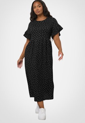 Kobieta ubrana w czarną sukienkę midi w białe kropki z falbankami na krótkich rękawach, w połączeniu z białymi trampkami, stojąca na białym tle.