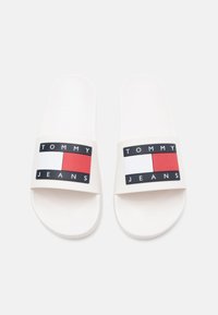 Valged liugjalatsid, mille rihmal on musta, punast ja valget värvi logo koos tekstiga "TOMMY JEANS"; sujuva ja tasase tallaga disain.