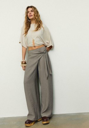 Femme portant un haut court beige ample, un pantalon gris à jambes larges taille haute avec ceinture, des chaussures compensées marron et des bracelets, debout contre un mur blanc.