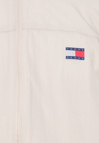 Šviesiai kreminės spalvos užtrauktukas striukė, pagaminta iš lygaus audinio. Kairėje pusėje yra stačiakampis mėlynas ir raudonas logotipo pleistras su užrašu "TOMMY JEANS".