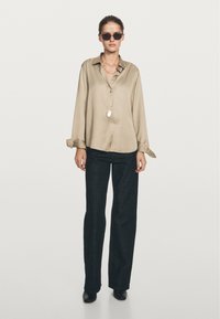 Woman wearing beige loose satin blouse, black wide-leg pants, black shoes, silver chain necklace with pendant, and dark sunglasses standing on white background.