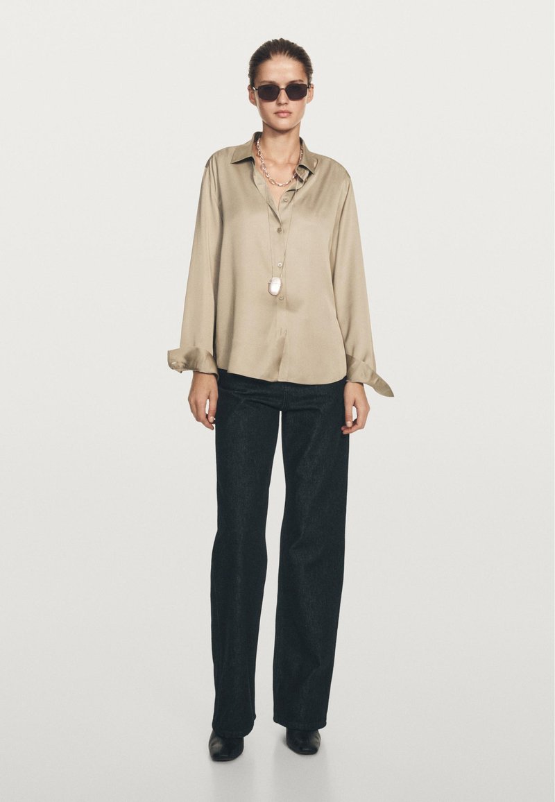 Woman wearing beige loose satin blouse, black wide-leg pants, black shoes, silver chain necklace with pendant, and dark sunglasses standing on white background.